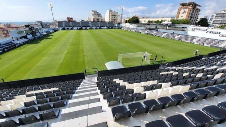 Vista desde las gradas con asientos negros y blancos, campo de juego y edificios urbanos en el horizonte