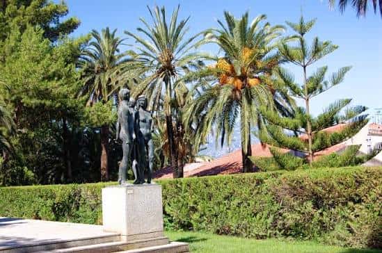 Escultura de figuras humanas en el jardín rodeada de palmeras y setos