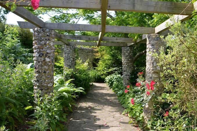 Paseo sombreado bajo un enrejado de madera con columnas de guijarros y flores rojas