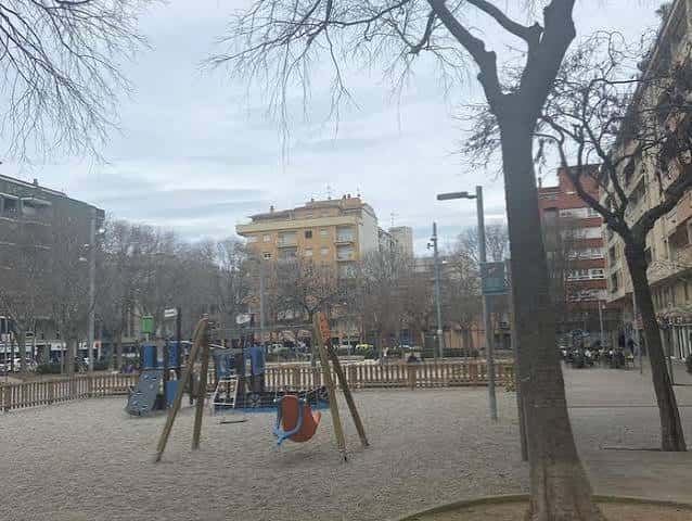 Área de juegos infantiles en la Plaça de Santa Eugènia con árboles y edificios al fondo