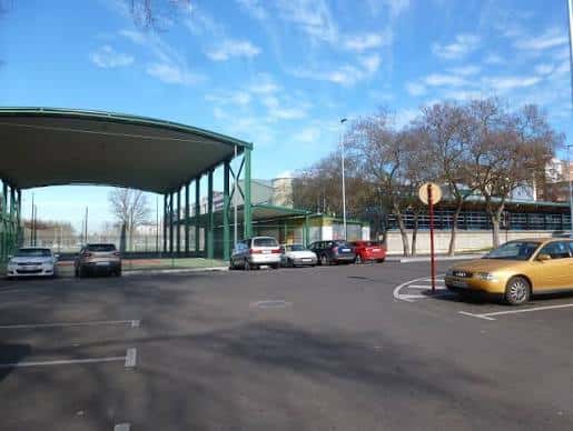 Estacionamiento frente al complejo deportivo con techado verde y vehículos aparcados