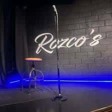 Escenario vacío de Rozco's Comedy Club con micrófono y taburete bajo luces azules