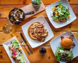 Platos de comida servidos en mesa de madera, incluyendo ensaladas y hamburguesas