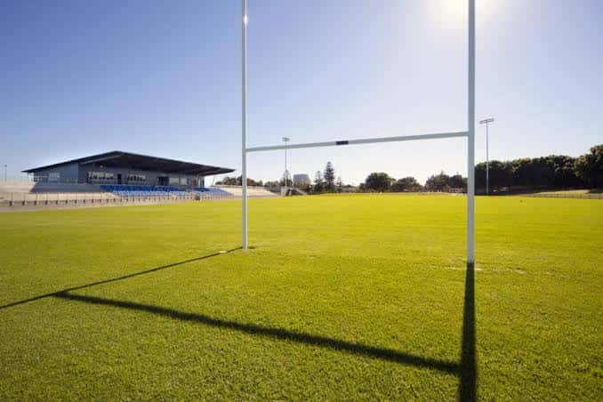 Campo de juego verde con portería de rugby y gradas al fondo