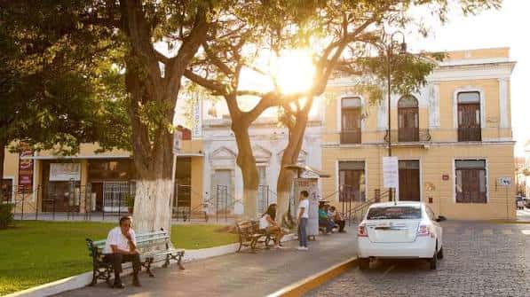 Atardecer en el parque con árboles frondosos y edificios coloniales al fondo