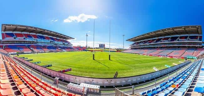 Vista interior del estadio mostrando las gradas rojas y azules, el campo de juego y los postes de rugby bajo el sol