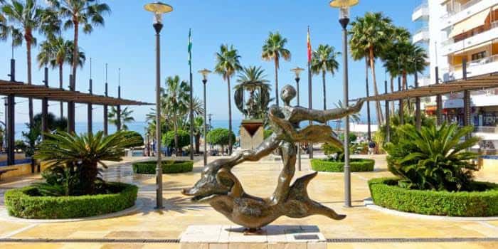 Escultura de delfines y figuras humanas en la Avenida del Mar con palmeras y mar al fondo