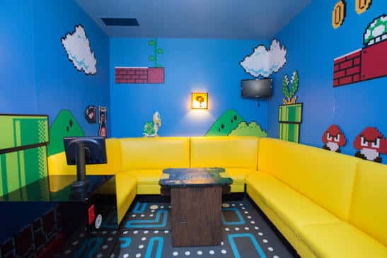 Sala de karaoke con temática de videojuegos retro, paredes azules y sofá amarillo