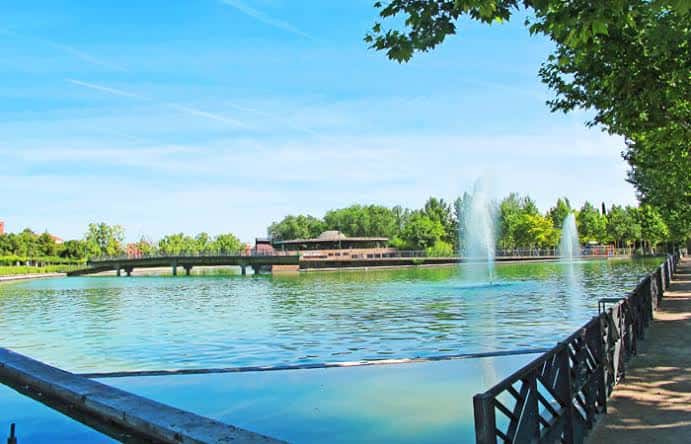 Paisaje del lago con puente peatonal, fuentes y zona de descanso arbolada