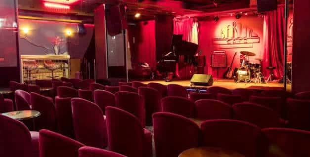 Vista del interior del club con escenario, piano y butacas rojas