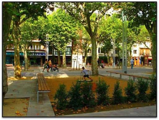 Plaza arbolada con personas caminando y bancos de madera