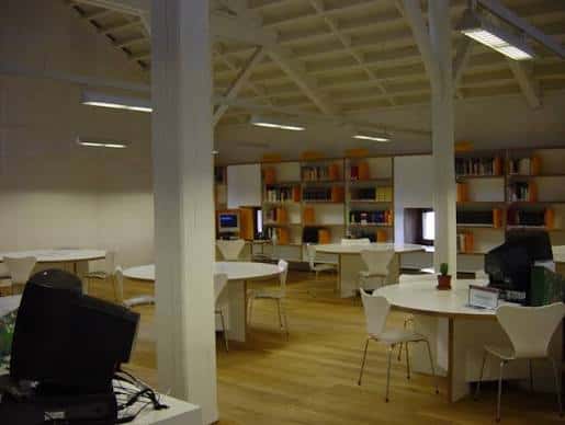 Interior de la biblioteca con mesas redondas blancas, sillas modernas y estanterías de libros en tonos naranjas
