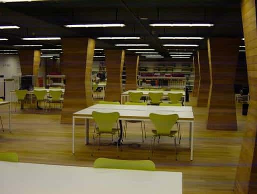 Sala de lectura con mesas largas, sillas verdes y columnas de madera curvadas bajo techo oscuro con iluminación lineal