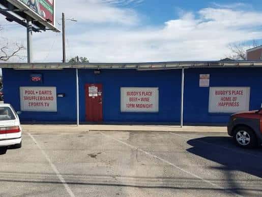 Fachada azul de Buddy's Place durante el día con carteles de 'Pool, Darts, Shuffleboard' y 'Home of Happiness'