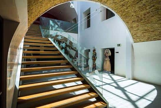 Escalera interior con barandilla de cristal y marionetas expuestas bajo arco de ladrillo