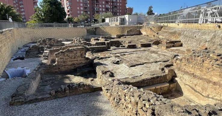 Área arqueológica con estructuras de piedra y edificios modernos al fondo