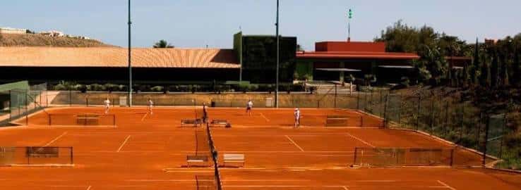 Pistas de tenis en uso con jugadores en acción y edificio principal del club al fondo
