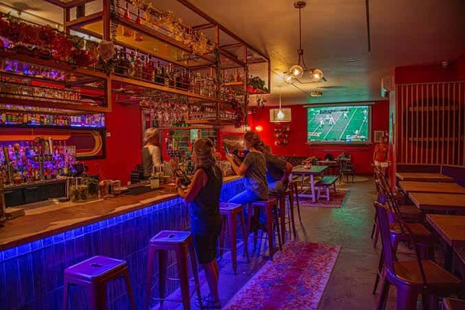 Interior del bar con estanterías de botellas, barra iluminada y clientes en los taburetes