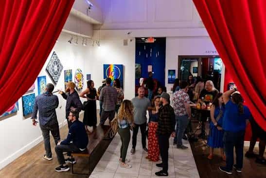 Interior de ArtUs Co mostrando una galería de arte con obras expuestas y una multitud de personas asistiendo a un evento social