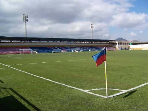 Campo de fútbol con portería, banderín de esquina y gradas bajo cielo nublado