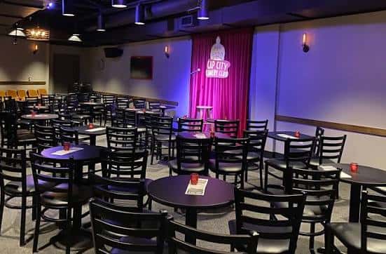 Interior del club con mesas, sillas y escenario con cortinas rojas y logotipo de Cap City Comedy Club