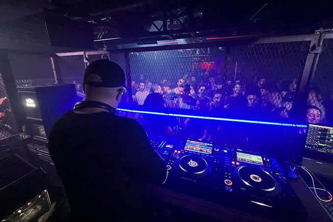 DJ actuando en el Club de Moog con luces azules y público detrás de una reja metálica
