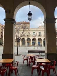 Interior de la plaza con arcos, mesas rojas y vista al edificio histórico al fondo