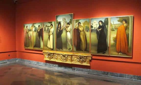 Sala de exposiciones con obras de Julio Romero de Torres enmarcadas en dorado sobre pared roja