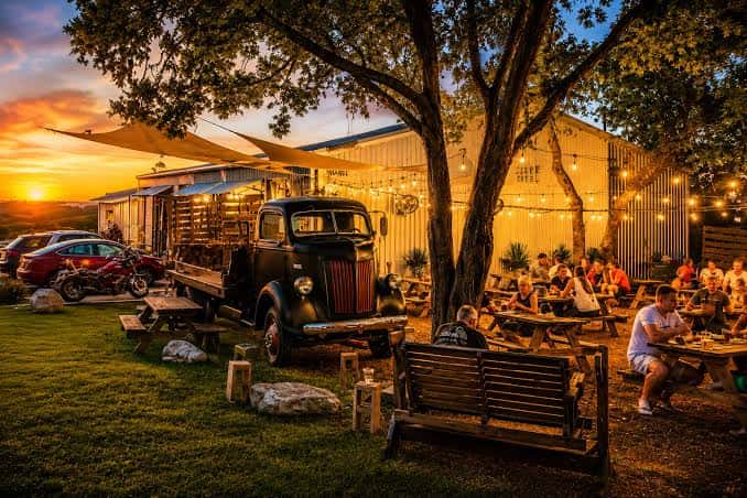 Vista exterior de Infamous Brewing Company al atardecer con clientes en mesas de picnic y decoración con luces