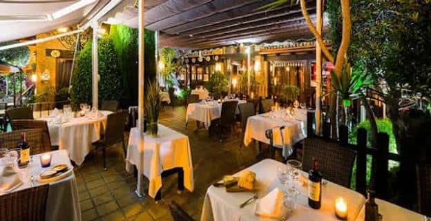 Interior al aire libre del restaurante con mesas cubiertas, iluminación cálida y entorno natural con árboles y plantas