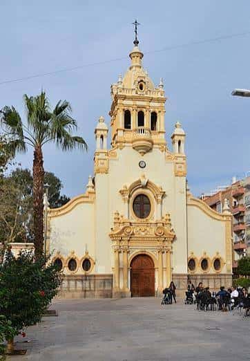 Vista frontal de la iglesia con palmera y personas sentadas en la plaza