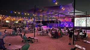 Vista nocturna del patio exterior de Drinks Backyard con luces de colores y mesas de picnic