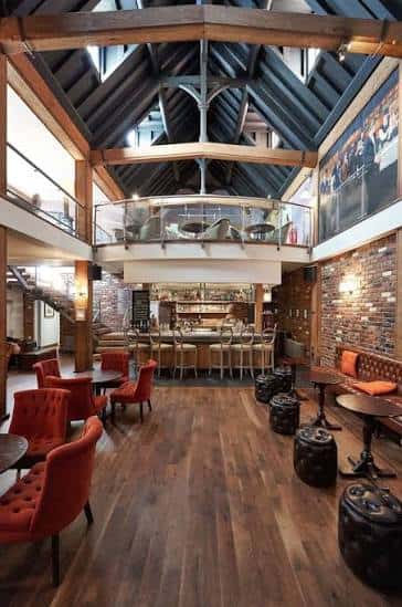Interior del bar del hotel con techo alto, vigas de madera y zona de estar con sofás rojos