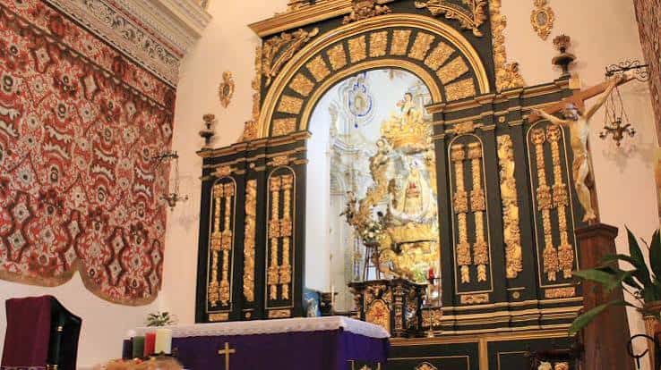 Interior del altar mayor con retablo dorado, cruz y tapices ornamentales en tonos rojos y dorados