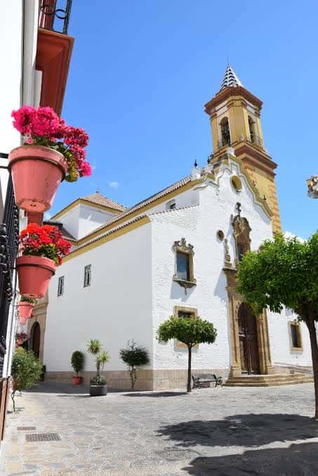 Fachada lateral de la iglesia con macetas de flores y árboles en la plaza, mostrando el entorno urbano