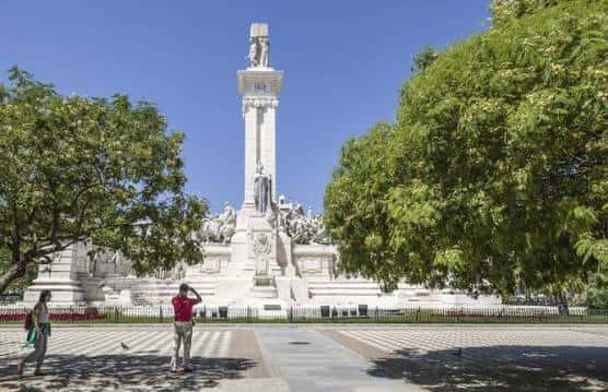 Plaza de España con el monumento al fondo y visitantes paseando entre árboles frondosos