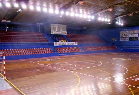 Pista de baloncesto interior con gradas rojas y azules, sin público, ideal para eventos deportivos o culturales