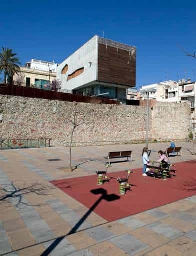 Plaza con zona de juegos infantiles y muro de piedra histórica al fondo