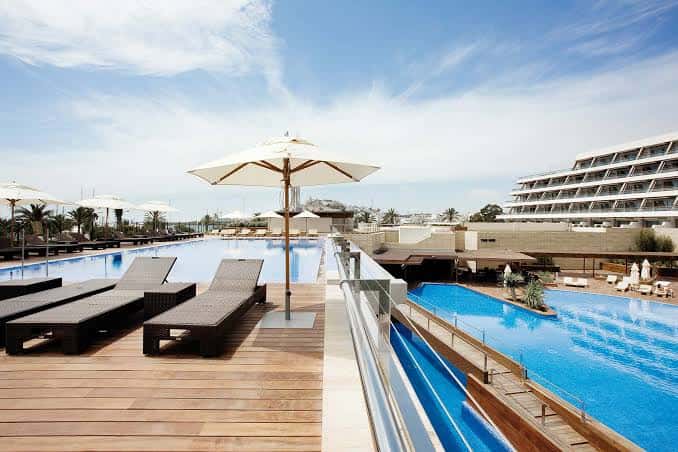 Terraza de piscina con tumbonas y sombrillas bajo cielo despejado, mostrando la arquitectura moderna del hotel