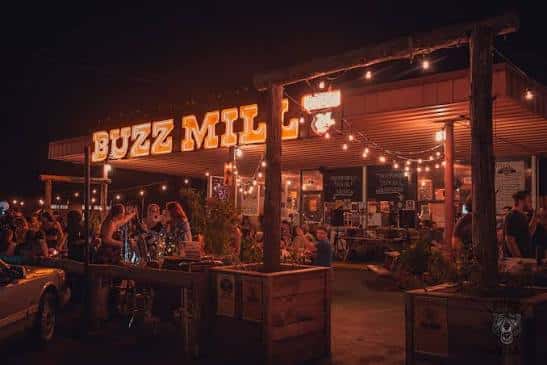 Vista nocturna del patio exterior del Buzz Mill con luces de cadena y clientes disfrutando