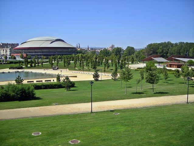 Amplia zona verde del parque con el Palacio de los Deportes en el horizonte