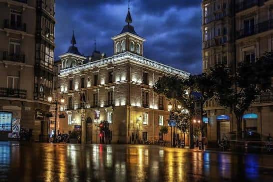 Vista nocturna del Palacio de los Chapiteles con fachada iluminada y reflejos en el suelo mojado
