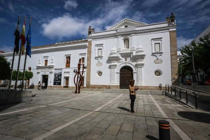 Fachada principal de la Asamblea de Extremadura con banderas y plaza peatonal frente al edificio