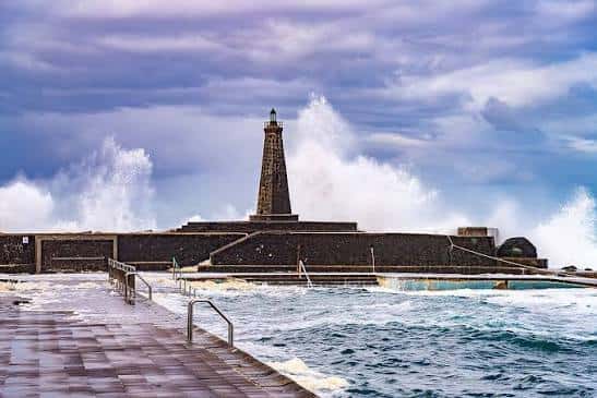 Faro de Bajamar con olas rompiendo contra el malecón y piscina de mar en primer plano