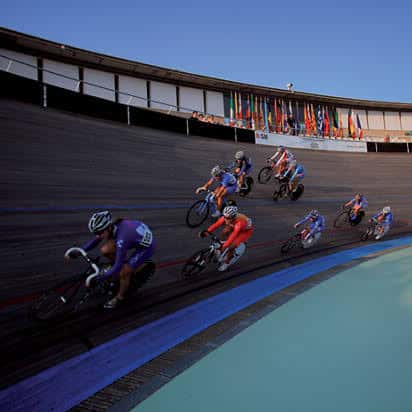 Ciclistas en competición sobre la pista de madera del velódromo, con banderas internacionales al fondo
