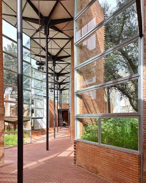 Pasillo interior con suelo de ladrillo, grandes ventanales y estructura metálica que conecta con el exterior