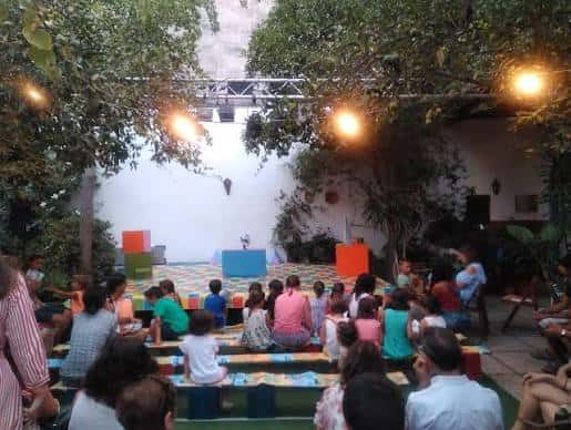 Vista general del patio de El Jardín de Berta durante un evento con público sentado en bancos y escenario al fondo