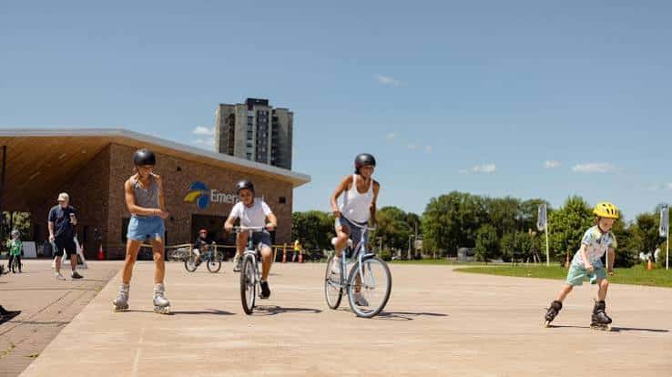 Niños patinando y en bicicleta frente a la entrada del Emera Oval en un día soleado