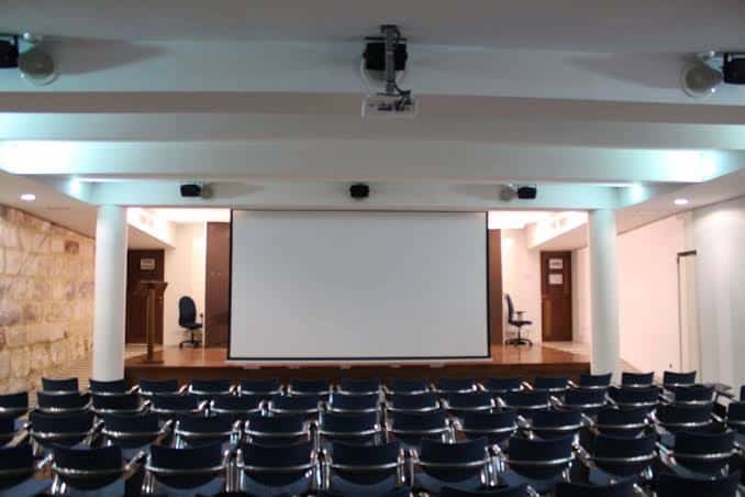 Sala de conferencias interior con proyector, pantalla y butacas dispuestas para eventos