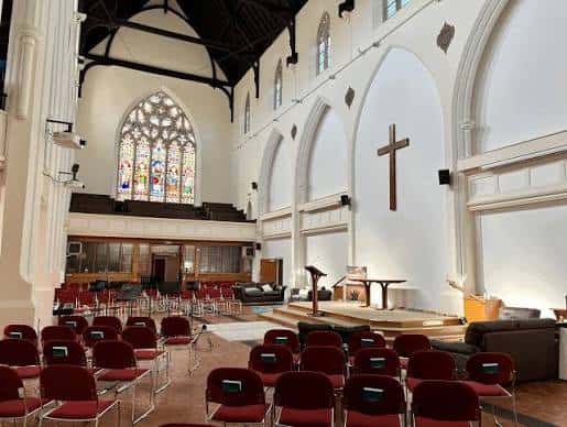 Interior del salón principal de la iglesia, configurado con sillas rojas para eventos, mostrando el altar y el vitral